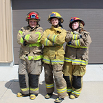 three teenage girls, dressed in firefighter uniforms and helmets, standing with crossed arms