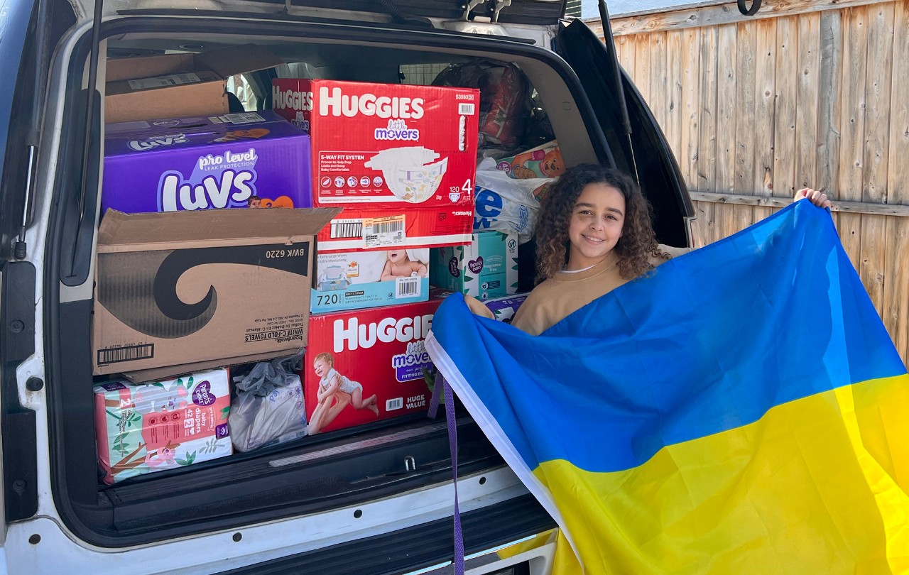 Girl Scout holding a Ukrainian flag in front of a SUV loaded with donations